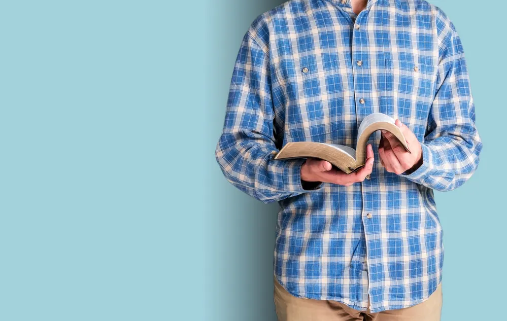 man standing against blue wall holding Bible (no face) for the post Five Biblical Reasons to be Silent and Blessings Related to Silence in the Bible